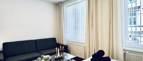 Hotelkamer met een bed, een zwarte bank, een glazen salontafel met kaarsen, en een stadszicht door grote ramen.