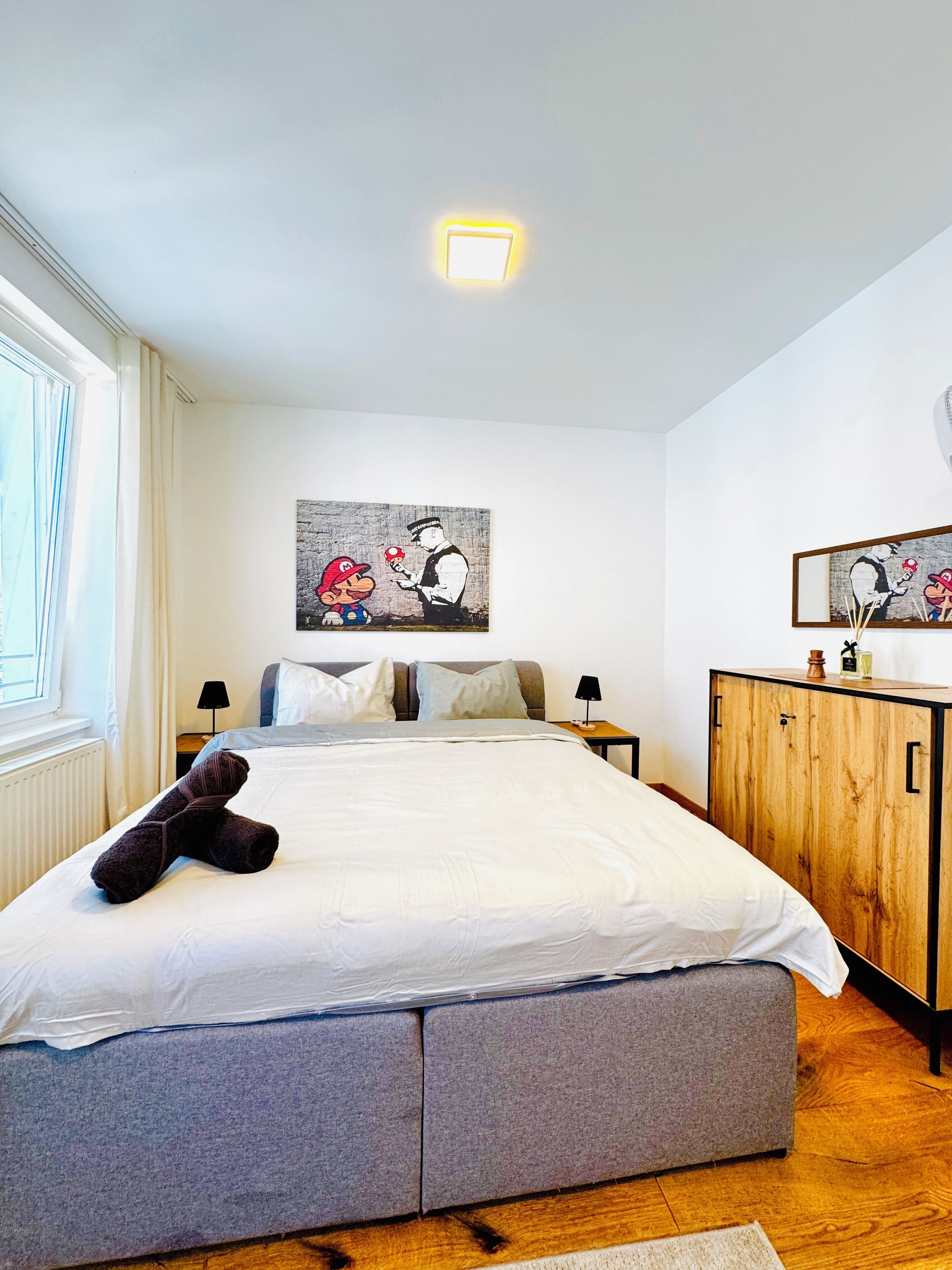A bedroom with a bed, a wooden wardrobe, a nightstand, a lamp, and a window.