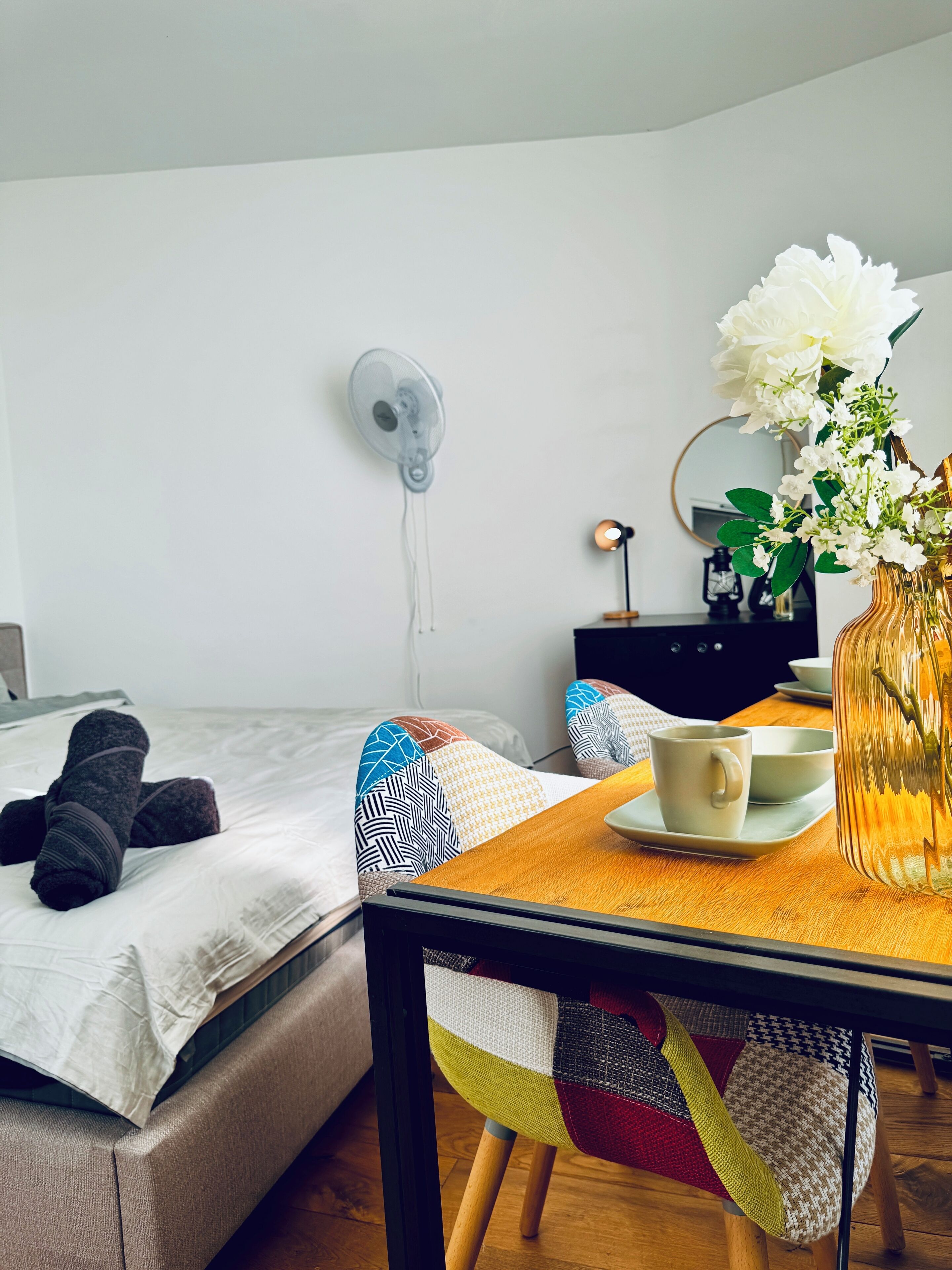 Cama bem arrumada com um edredom branco e um par de meias enroladas. Uma mesa de madeira com uma caneca branca e uma tigela pequena está em frente à cama. Um vaso com flores brancas está sobre a mesa.