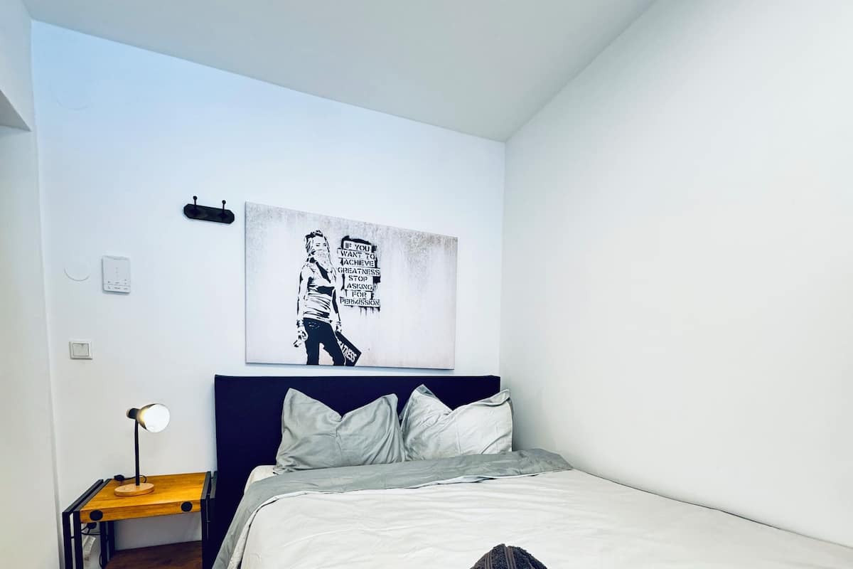 A bedroom with a bed, a bedside table with a lamp, a wall-mounted artwork, and a wooden floor.