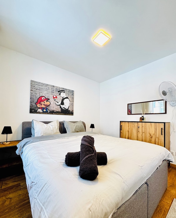 A bedroom with a bed, a headboard, a bedside table with a lamp, a wall-mounted mirror, a wooden cabinet, and a ceiling light fixture.