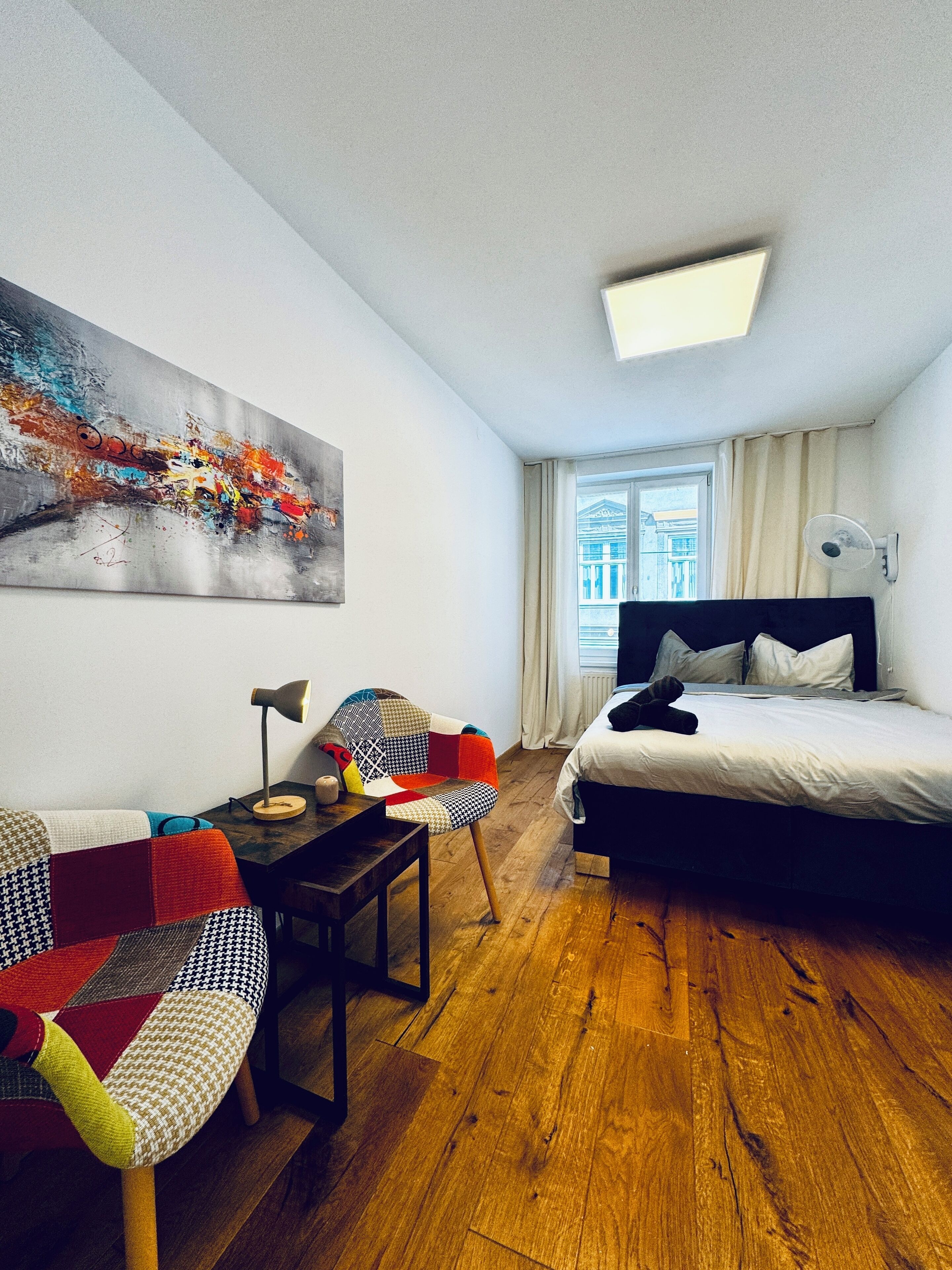 A modern hotel room with a large bed, a desk with a lamp, a colorful chair, and a painting on the wall.