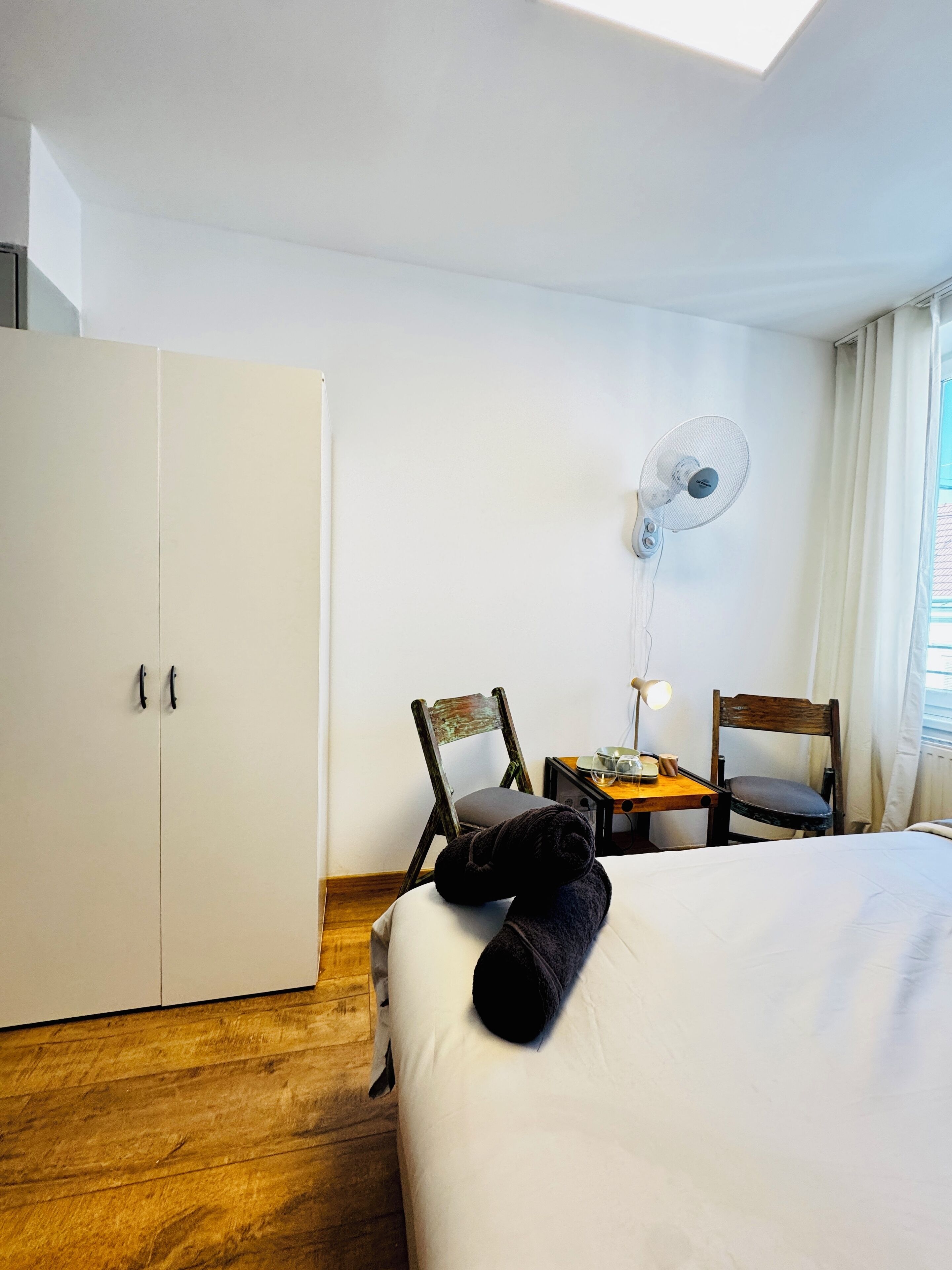 Ett hotellrum med en säng, ett skrivbord med en lampa, två stolar, en garderob och en takfläkt.