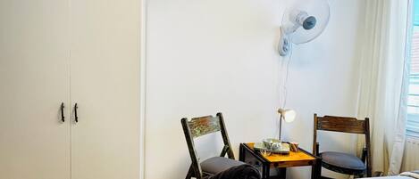 A hotel room with a bed, a desk with a lamp, two chairs, a wardrobe, and a ceiling fan.