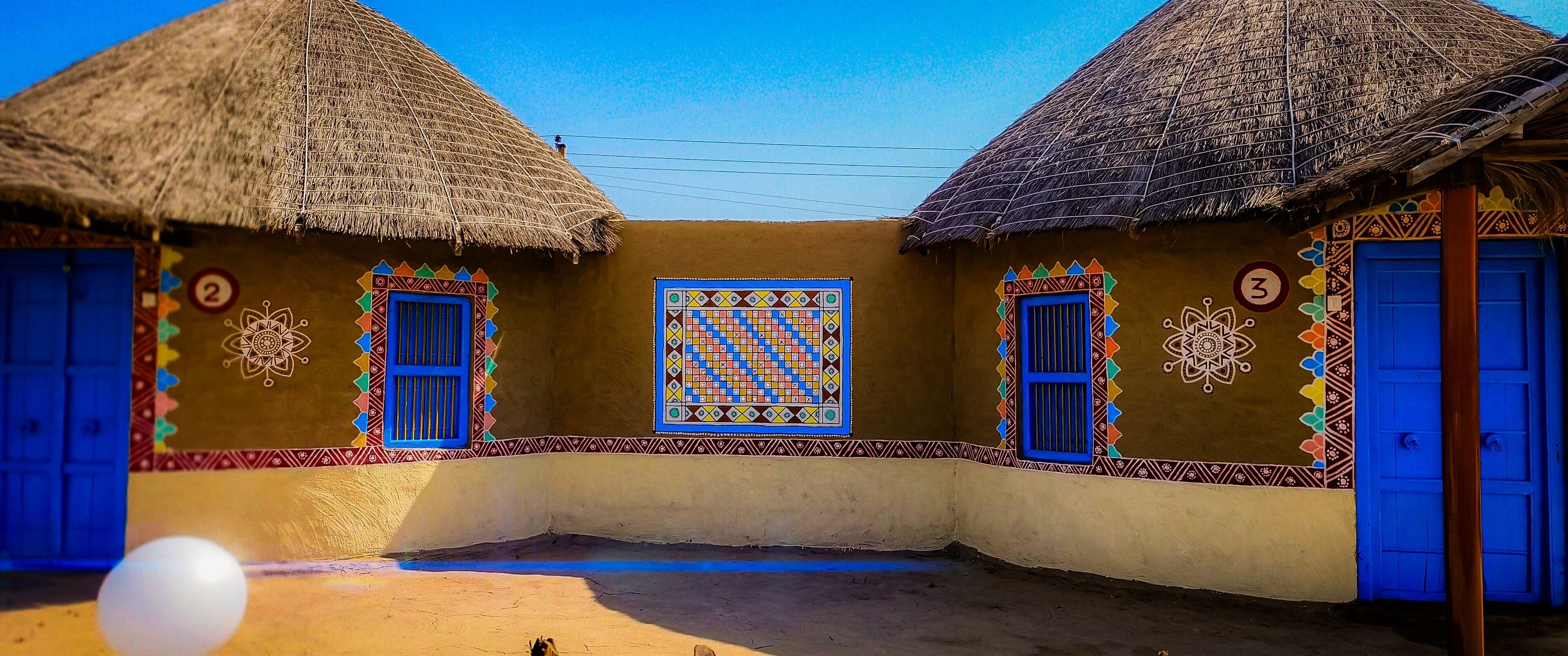 Un patio con dos edificios de techo de paja, cada uno con una puerta azul y una ventana con un diseño geométrico colorido.