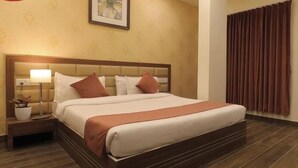 A hotel room with a large bed, a nightstand with a lamp, a potted plant, and a window with curtains. - Hotel Shiv Shakti  (Rajkot)