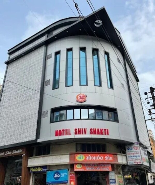 A hotel named "Hotel Shiv Shakti" with a modern architectural design, featuring a prominent sign and a storefront with various shops below. - Hotel Shiv Shakti  (Rajkot)