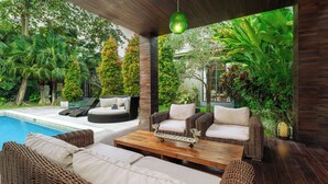 A poolside area with wicker furniture, a wooden table, and lush greenery. - Exclusive 5-Bedroom Luxury Villa in Canggu, Walking Distance to the Beach! (Tanggu)