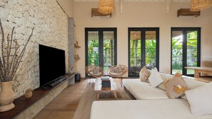 A living room with a stone wall, a flat-screen TV, wicker chairs, a white sofa, and large windows with greenery outside. - Exclusive 5-Bedroom Luxury Villa in Canggu, Walking Distance to the Beach! (Tanggu)