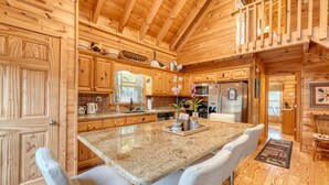 A wooden cabin interior with a large kitchen island, wooden cabinets, and a dining area with white cushioned chairs.