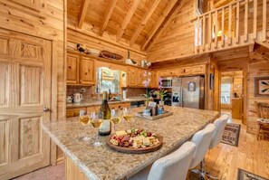 A wooden cabin interior with a kitchen area featuring granite countertops, a dining table with white chairs, and a buffet-style food arrangement. - Mountain Bearadise - Mountain views and privacy! 3 BR, 2 BA Cabin (Blairsville)