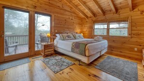 A wooden cabin bedroom with a bed, a nightstand, a window, and a door leading to an outdoor deck.