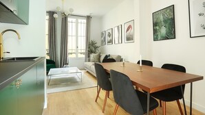 Un salon moderne avec une table à manger en bois, des chaises noires, un canapé et un coin cuisine doté d’un meuble vert et d’un robinet doré.