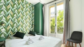 Une chambre d’hôtel avec un lit, des oreillers, une chaise, une fenêtre avec des rideaux et un mur vert orné d’un motif de feuilles.