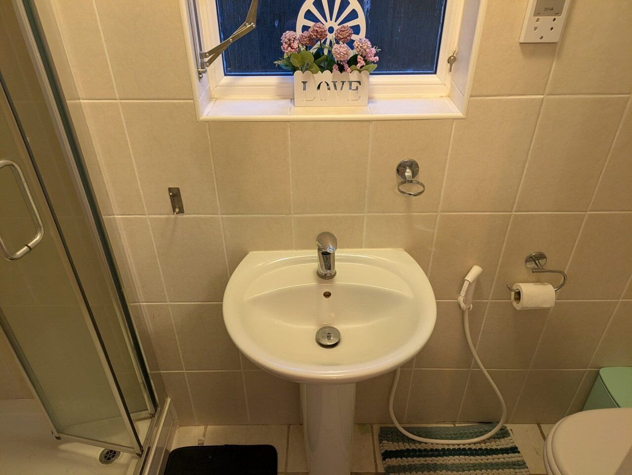 Un baño con un lavamanos blanco, una ventana con un cartel decorativo y una zona de ducha.