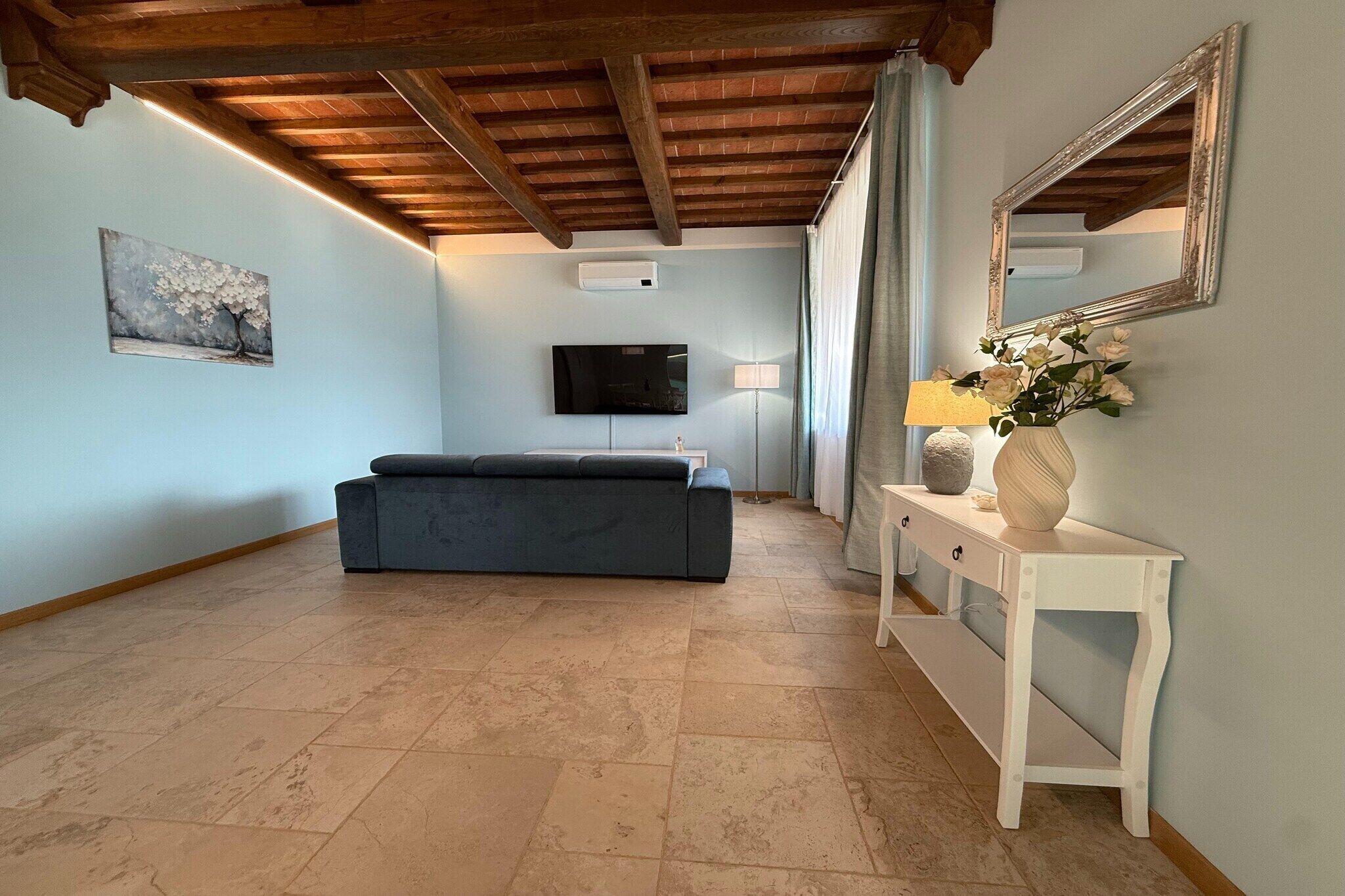 Una habitación con sofá, televisor montado en la pared, lámpara y un florero con flores sobre una consola.