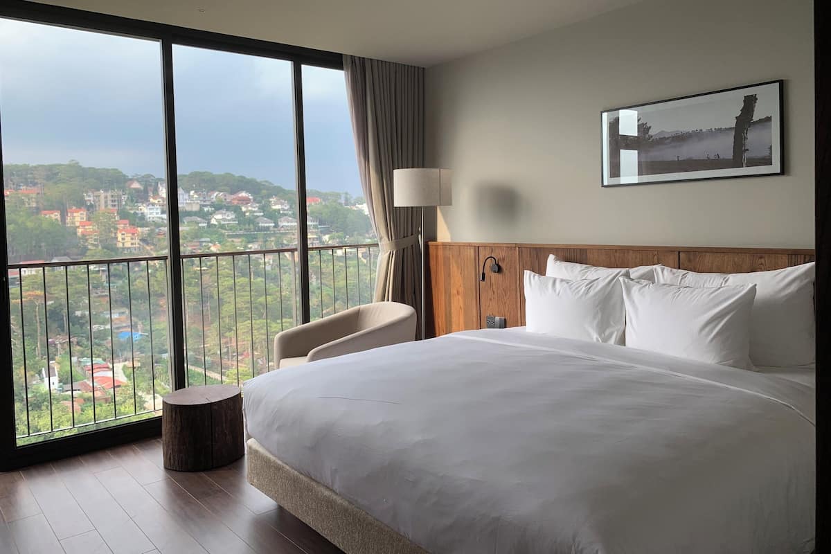 Habitación de hotel con una cama grande, una silla, una mesita de noche, una lámpara y un amplio ventanal con vistas a un paisaje urbano.