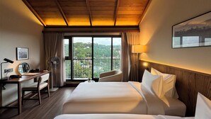 Een hotelkamer met een houten plafond, een groot bed, een bureau met stoel, een ventilator en uitzicht op een bergachtig landschap door het raam.