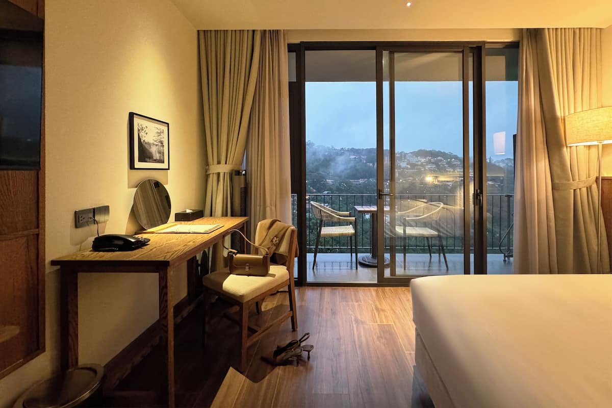 Habitación de hotel con escritorio, silla y cama. Se observa una vista panorámica de la ciudad a través de la ventana.