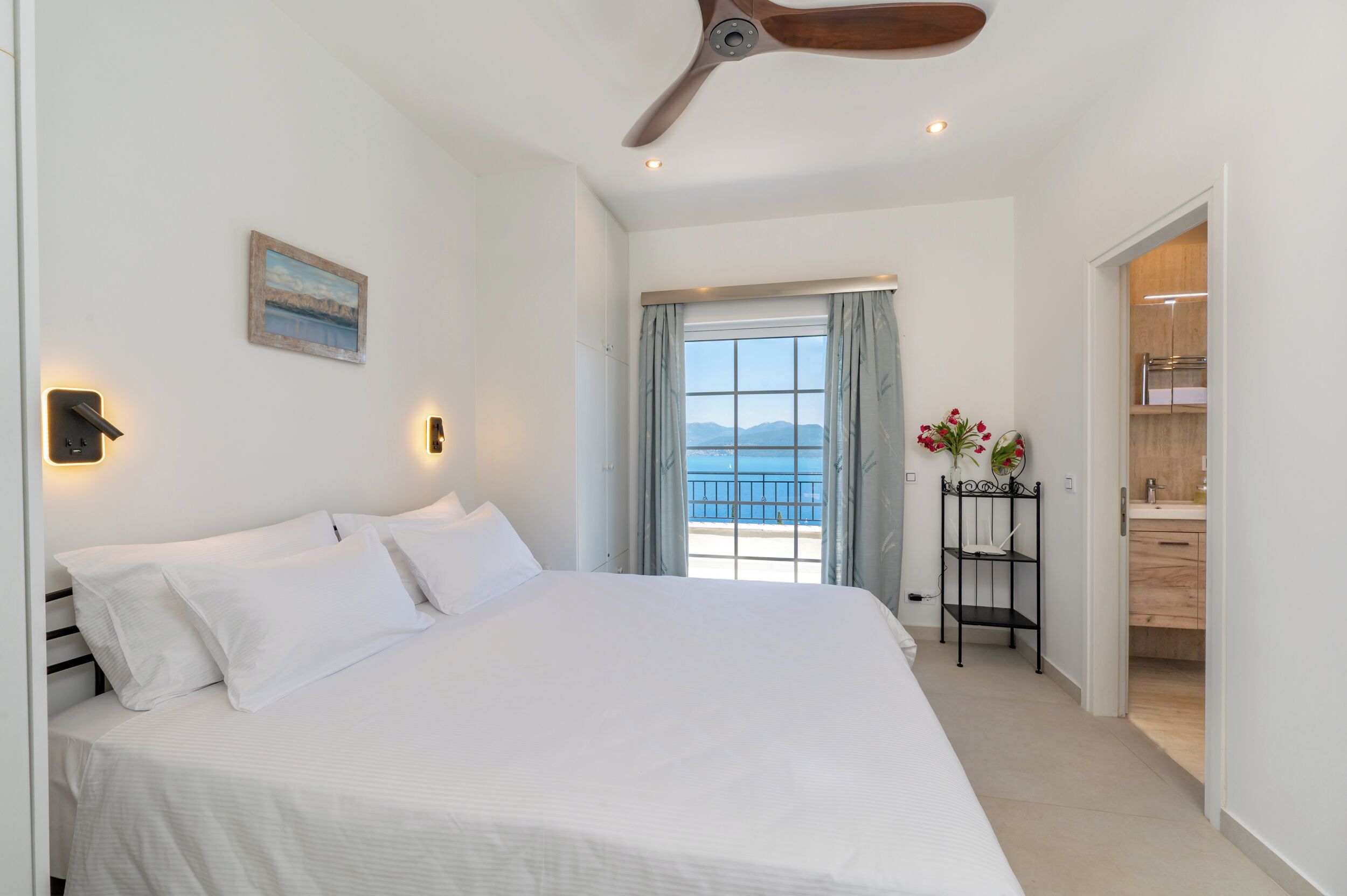 A bedroom with a large bed, a ceiling fan, a window with a view of the sea, and a bathroom with wooden cabinets.