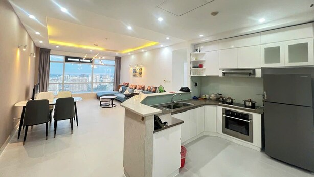 A modern kitchen with white cabinets, a built-in oven, and a refrigerator. There is a dining area with a table and chairs, and a sofa. - Victory Tower (Ho Chi Minh City)