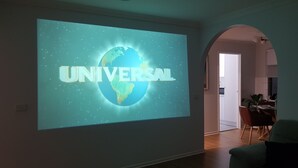 Ett vardagsrum med en stor skärm som visar Universal-logotypen, ett trägolv, en grön soffa och en matplats i bakgrunden.