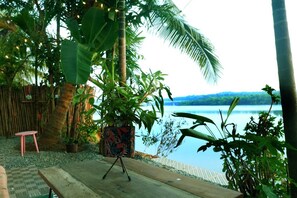 A wooden deck with a small table and a chair, surrounded by tropical plants and a view of a lake. - Riverside Sanctuary (Canacona)