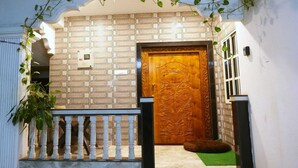 Une entrée d’immeuble avec un mur recouvert de carreaux, une porte en bois ornée de sculptures complexes, un petit tapis vert et une balustrade blanche.