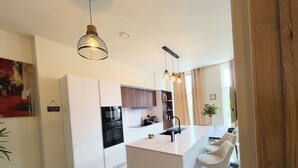 Una cuina moderna amb armaris blancs, una illa central amb tamborets, il·luminació penjant i vista a l'exterior.