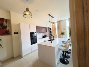 Een moderne keuken met witte kastjes, een centraal eiland met barkrukken, hanglampen en uitzicht op de omgeving.