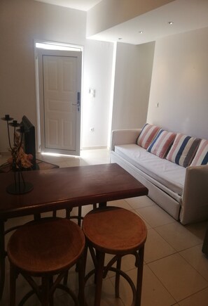 A living room with a sofa, a dining table with two stools, a door, and a small table with a vase. - One Bedroom Apartment with Private Patio - Suli Residences (Kefalonia)