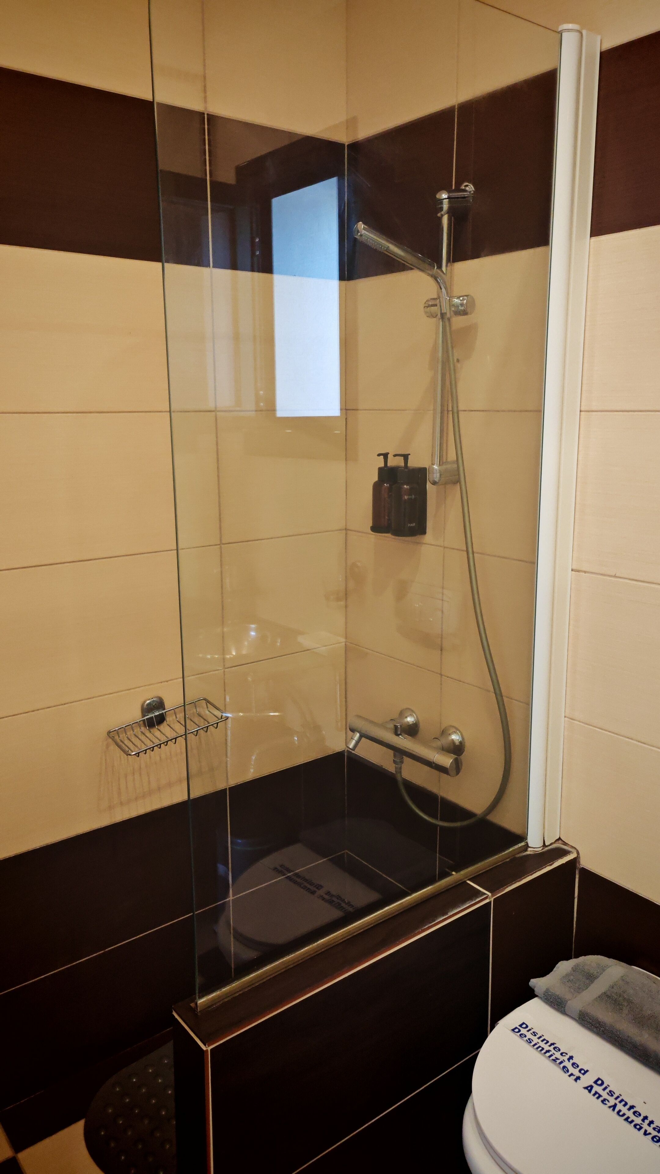A glass-enclosed shower stall with a handheld showerhead, a towel rack, and a disinfection notice on the toilet.