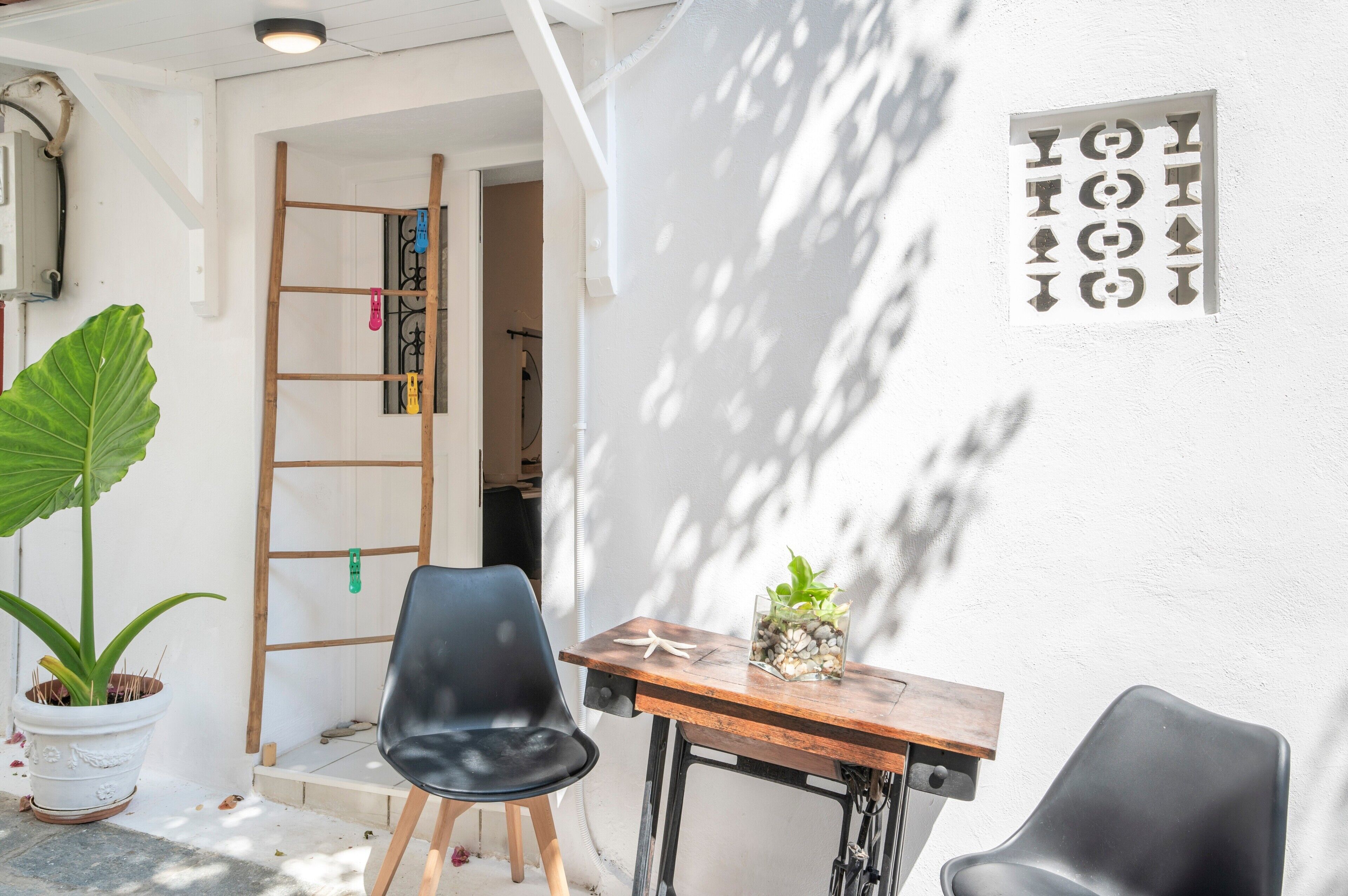 Een klein terras met een houten tafel en stoelen, een plant in pot, en een ladder die tegen een muur staat.