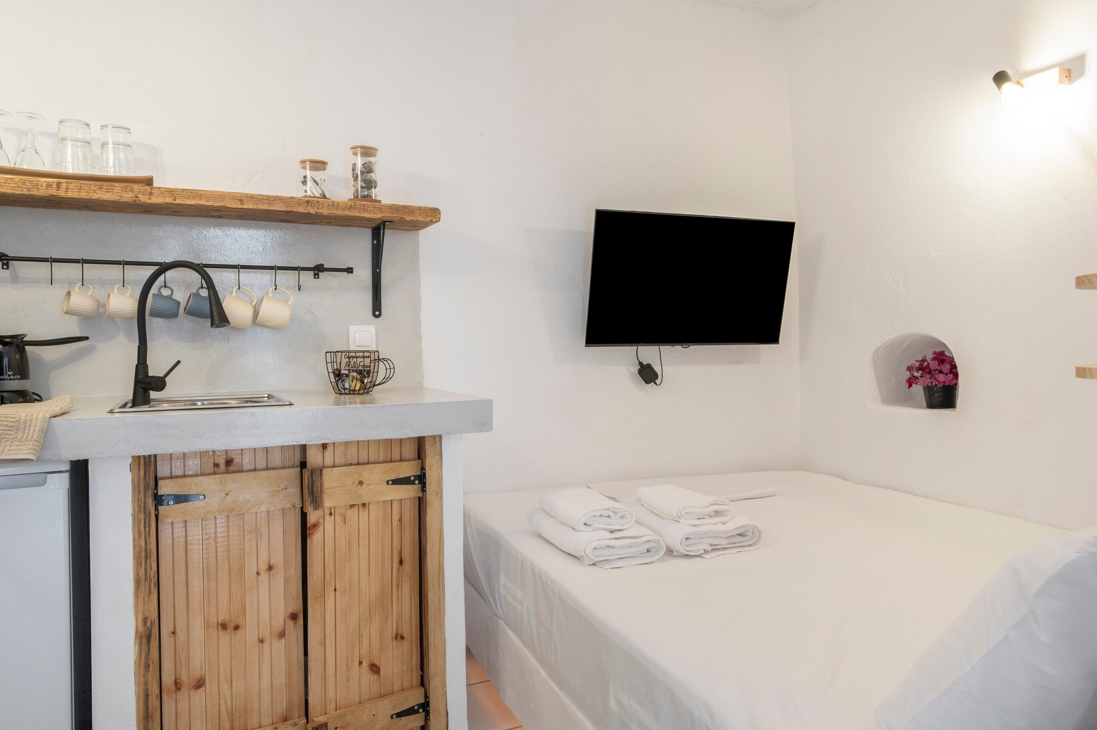 Een compacte kamer met kitchenette, een bed met netjes opgevouwen handdoeken, een wandtelevisie en een klein plankje met decoratieve objecten.