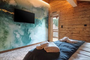 A room with a wooden ceiling, a flat-screen TV mounted on the wall, a bed with a blue mattress and white pillows, and a window with a view of trees. - Chalet Phoenix - 14 guests - Spa & Hammam, Gérardmer (Gérardmer)