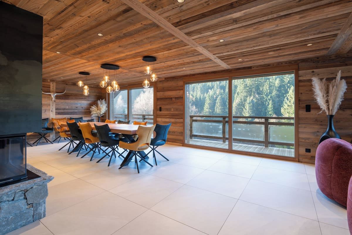 A spacious dining area with a large table and chairs, wooden ceiling, and large windows with a forest view.