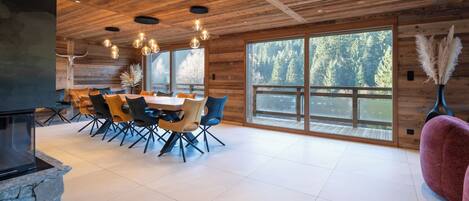 A spacious dining area with a large table and chairs, wooden ceiling, and large windows with a forest view.