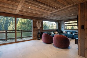 A spacious room with a large window, a dark-colored sofa, and two round red ottomans. - Chalet Phoenix - 14 guests - Spa & Hammam, Gérardmer (Gérardmer)