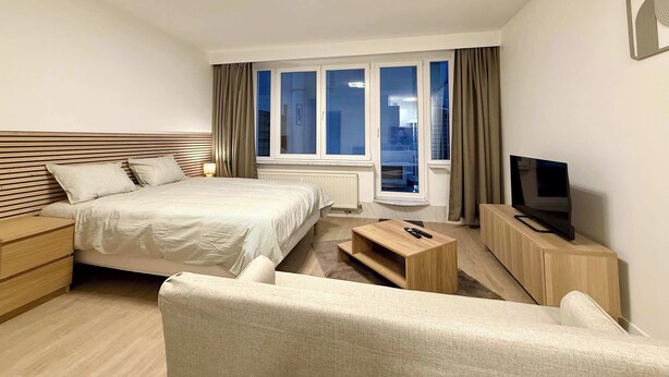 A modern hotel room with a bed, a TV, a wooden dresser, and a window with curtains. - Studio moderne au coeur du quartier Saint Adalbert (Liège)