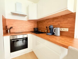 Une cuisine compacte avec des meubles blancs, un plan de travail en bois, un four encastré et une machine à café.