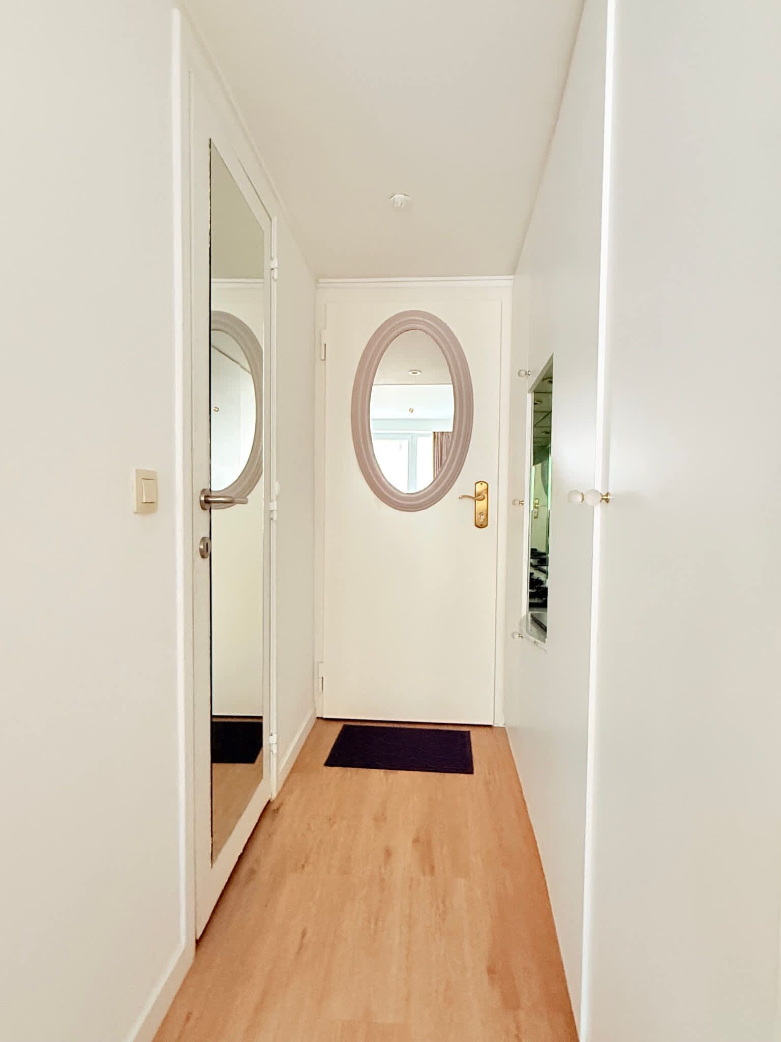Een gang met een houten vloer, witte wanden en een deur met een ronde spiegel.