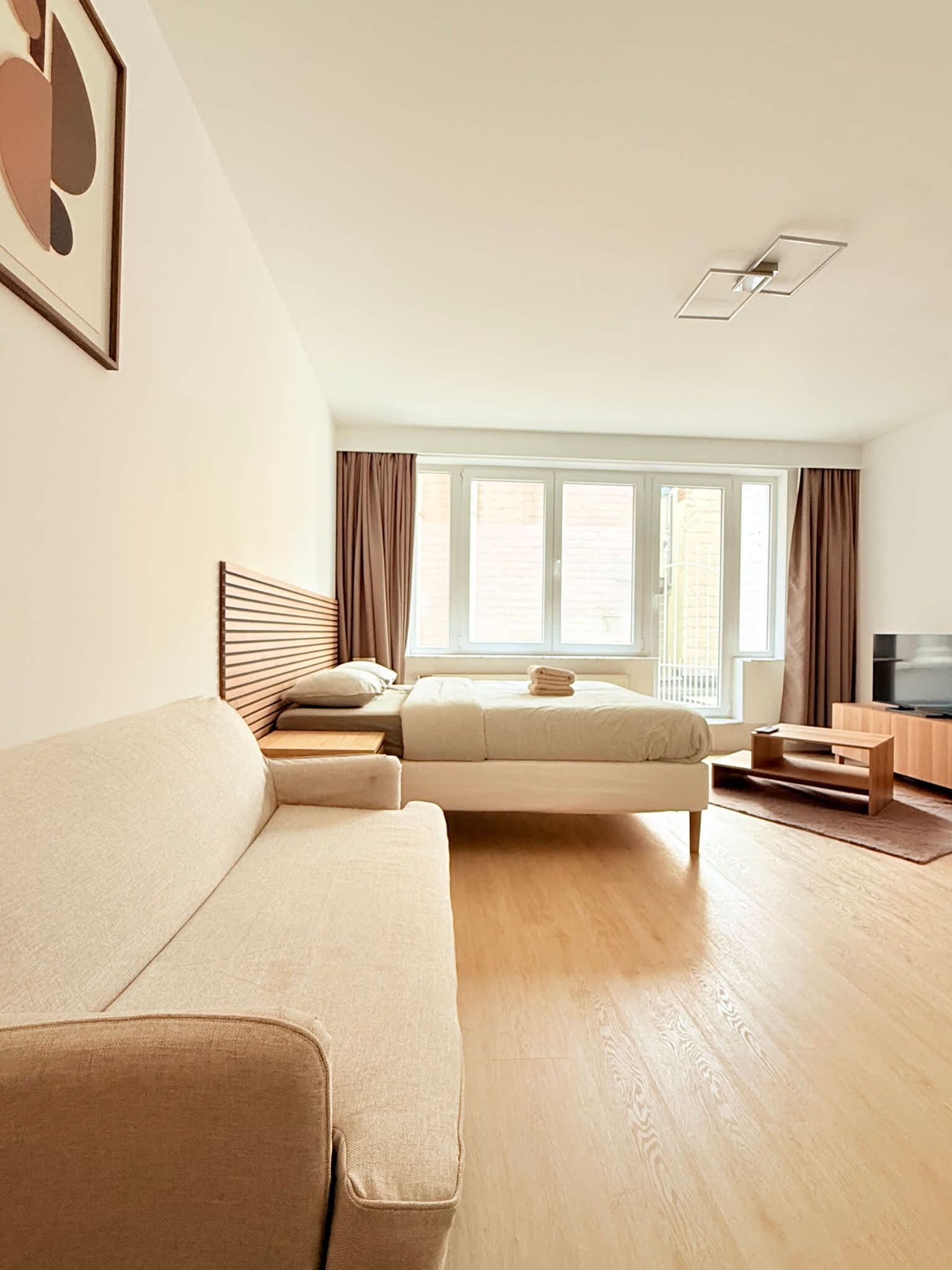Een moderne woonkamer met een bank, een bed, een televisie en een groot raam met gordijnen.