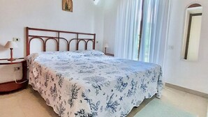 A bedroom with a wooden bed frame, a floral bedspread, a nightstand with a lamp, a wall-mounted picture, and a window with white curtains. - <b></b>Located on the southeastern coast of Sardinia, 20 km from Villasimius, <b>Villa Venere</b> is situated approximately 2 km from the center of Costa Rei. Surrounded by a spacious and well-kept private garden of about 200 sqm, this charming villa is  (Costa Rei)