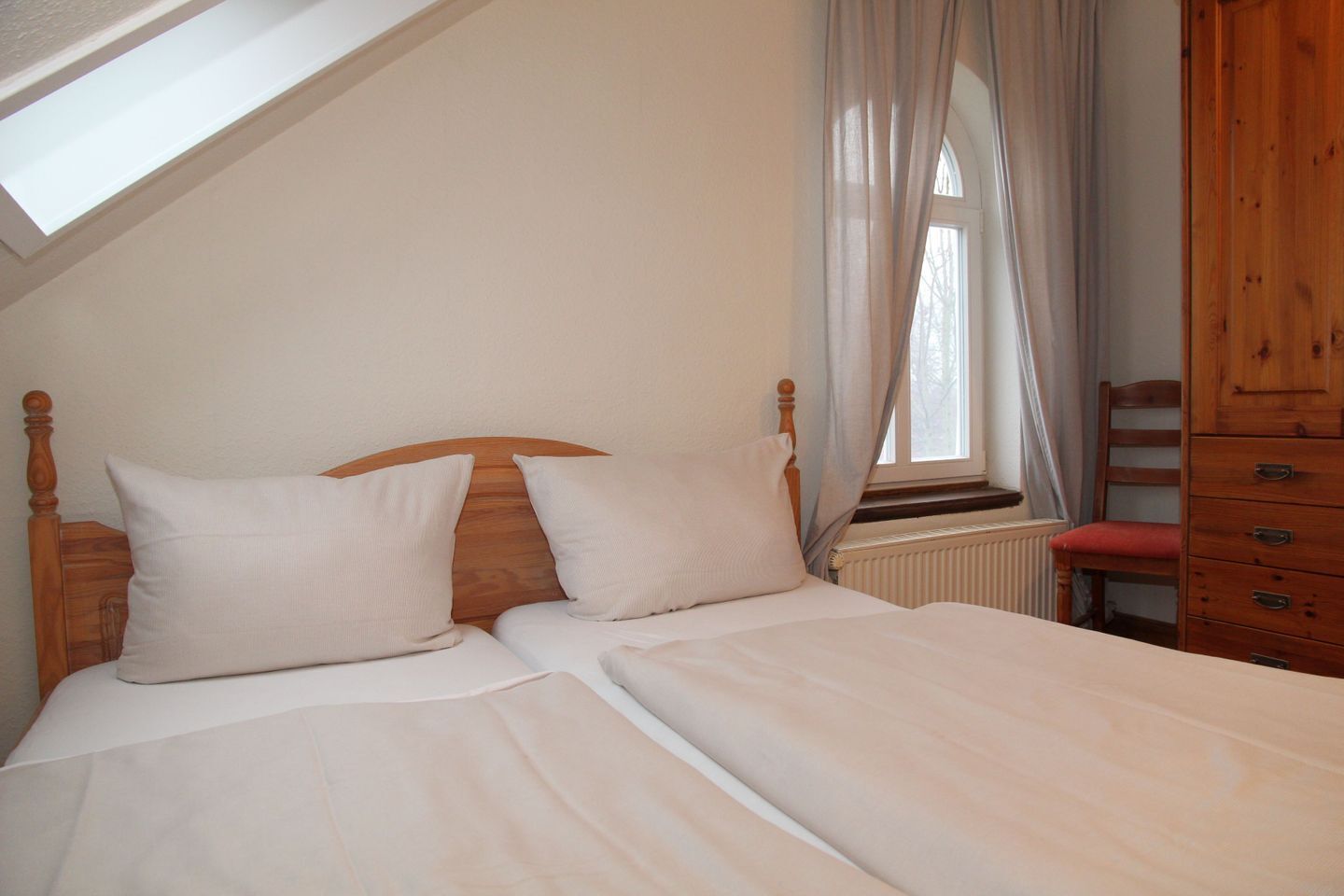 Un dormitorio con techo inclinado, estructura de cama de madera, dos almohadas, un radiador, un armario de madera y una ventana con cortinas traslúcidas.