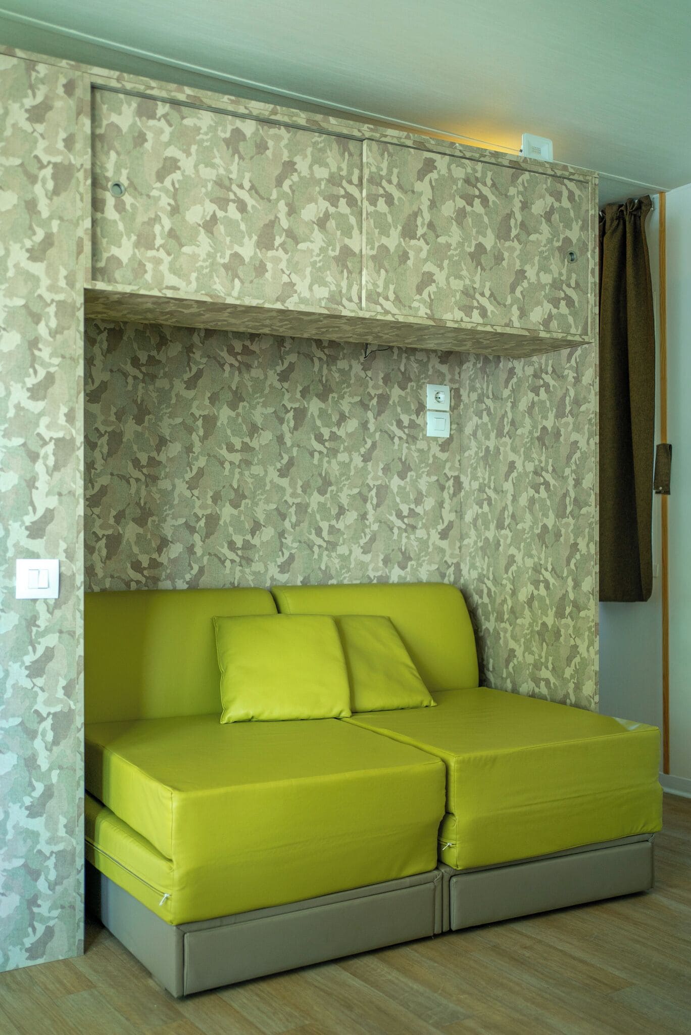 A room with a camouflage-patterned wall, a bright yellow sofa, and a brown curtain.
