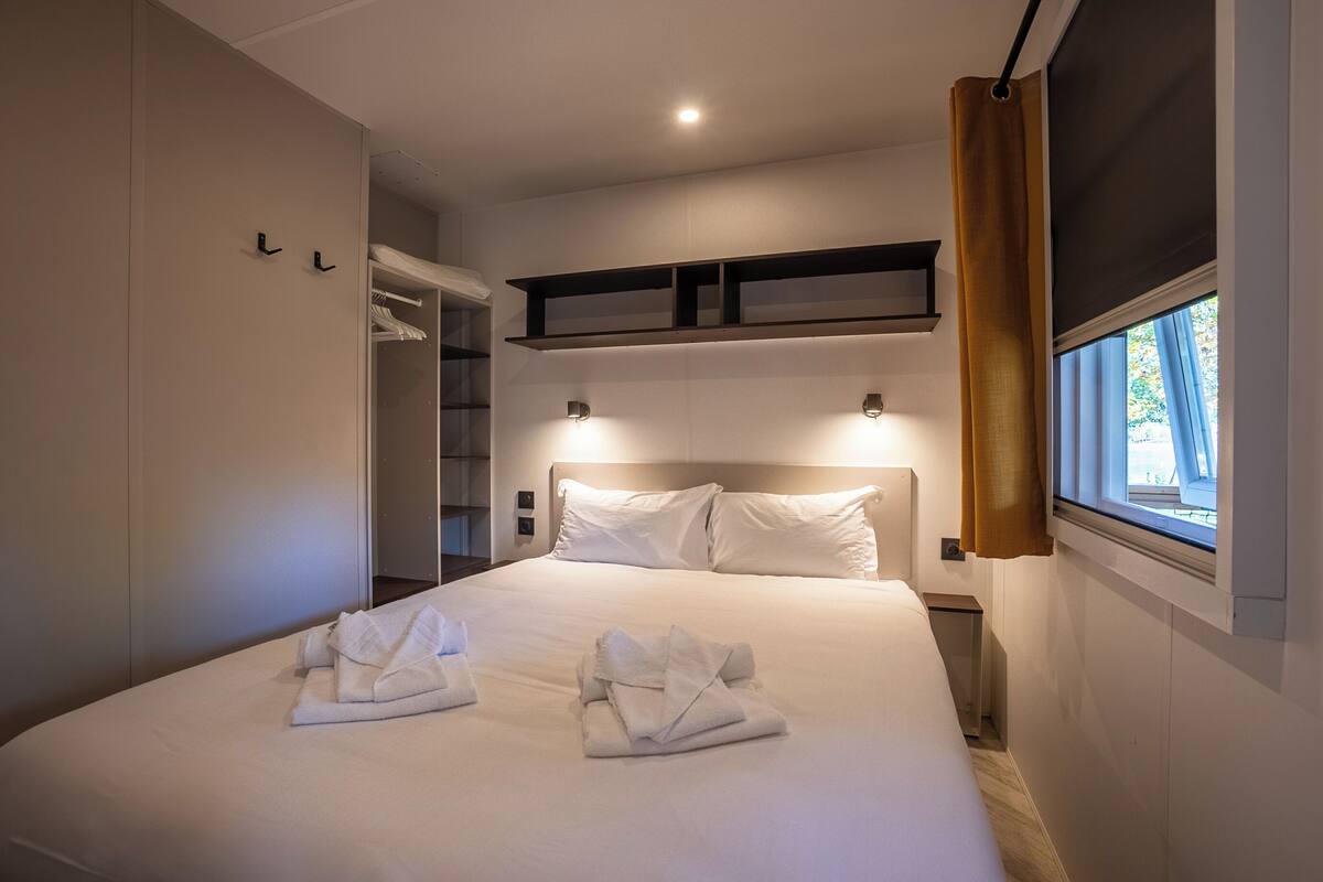 Una habitación de hotel con una cama grande, un armario, un estante sobre la cama y una ventana con cortinas.