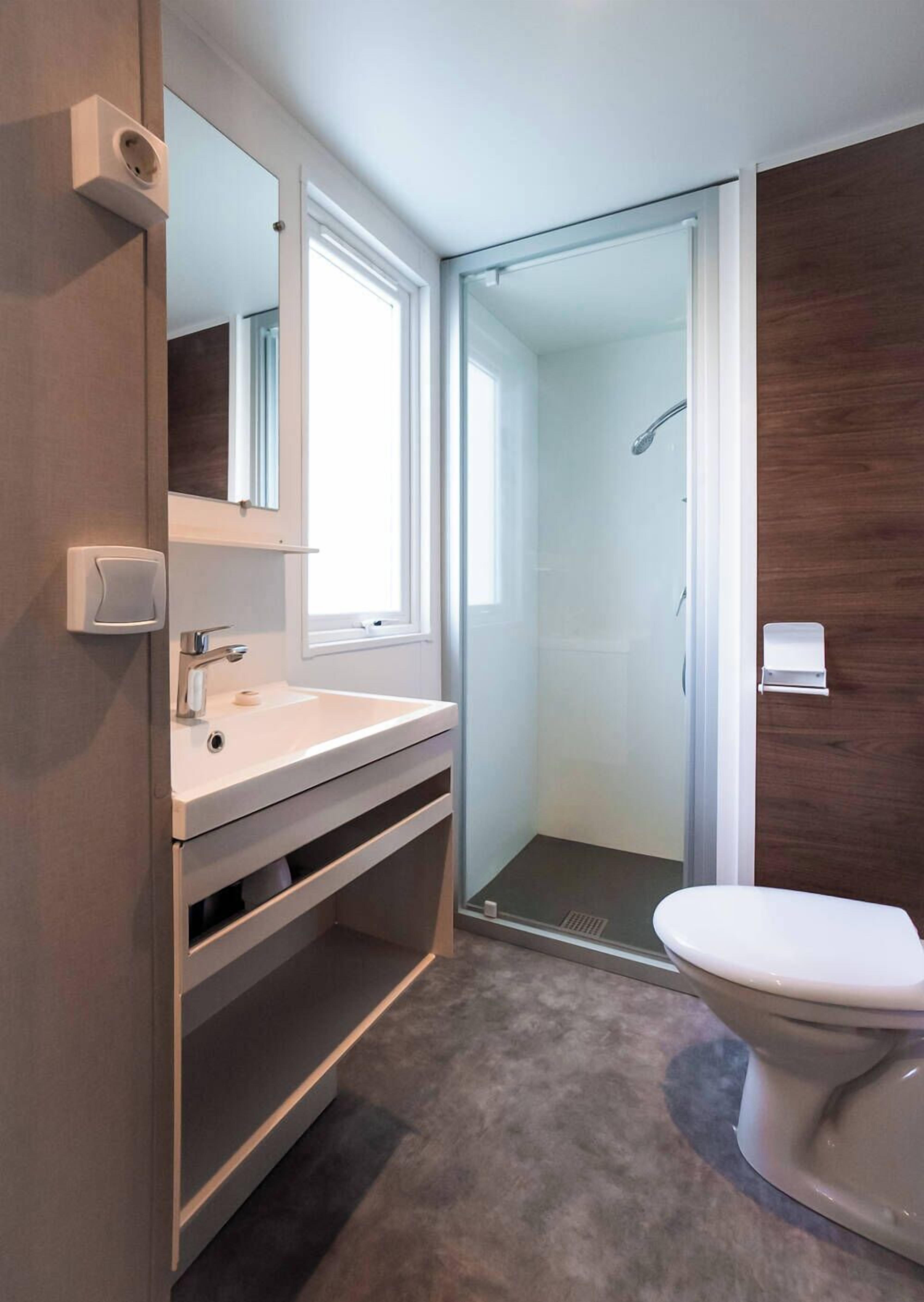 Une salle de bain moderne avec un lavabo blanc, un miroir et des toilettes. Elle comprend un espace de douche vitré et un panneau mural en bois.