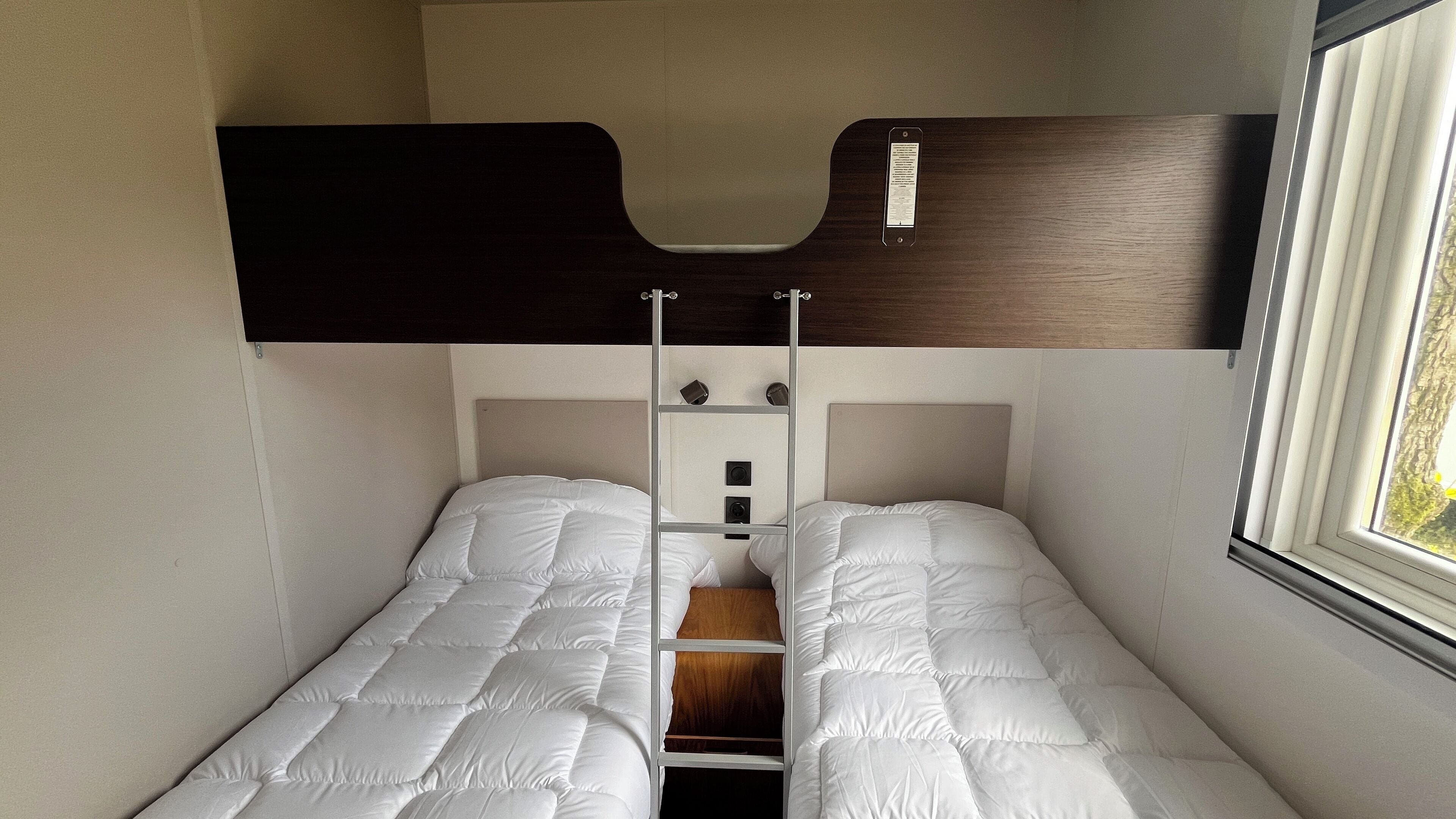 A small, compact room with two single beds, a wooden headboard, and a window providing natural light.