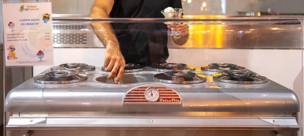 Une personne se tient derrière un distributeur de crème glacée en acier inoxydable, doté de plusieurs compartiments et portant l’inscription « Dolce Vita ».