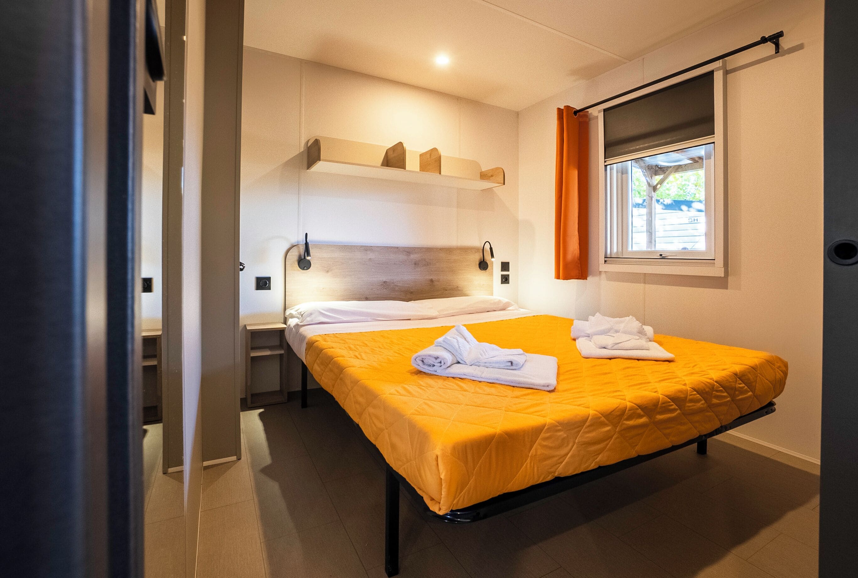A neatly made bed with a yellow bedspread and white linens, a wooden headboard, and a window with orange curtains.